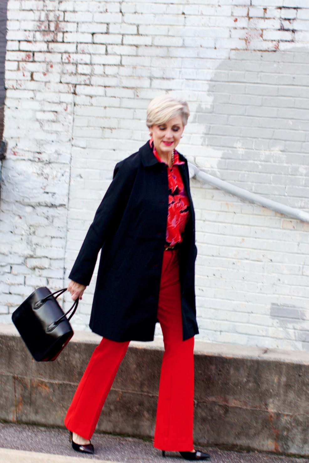 beth from Style at a Certain Age wears Anthropologie red pants, pink and red shirt, Everlane black raincoat, and black suede pumps