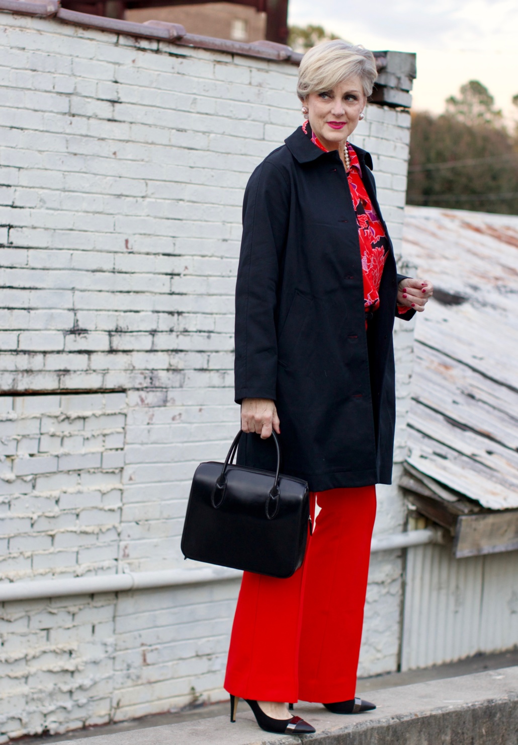 beth from Style at a Certain Age wears Anthropologie red pants, pink and red shirt, Everlane black raincoat, and black suede pumps