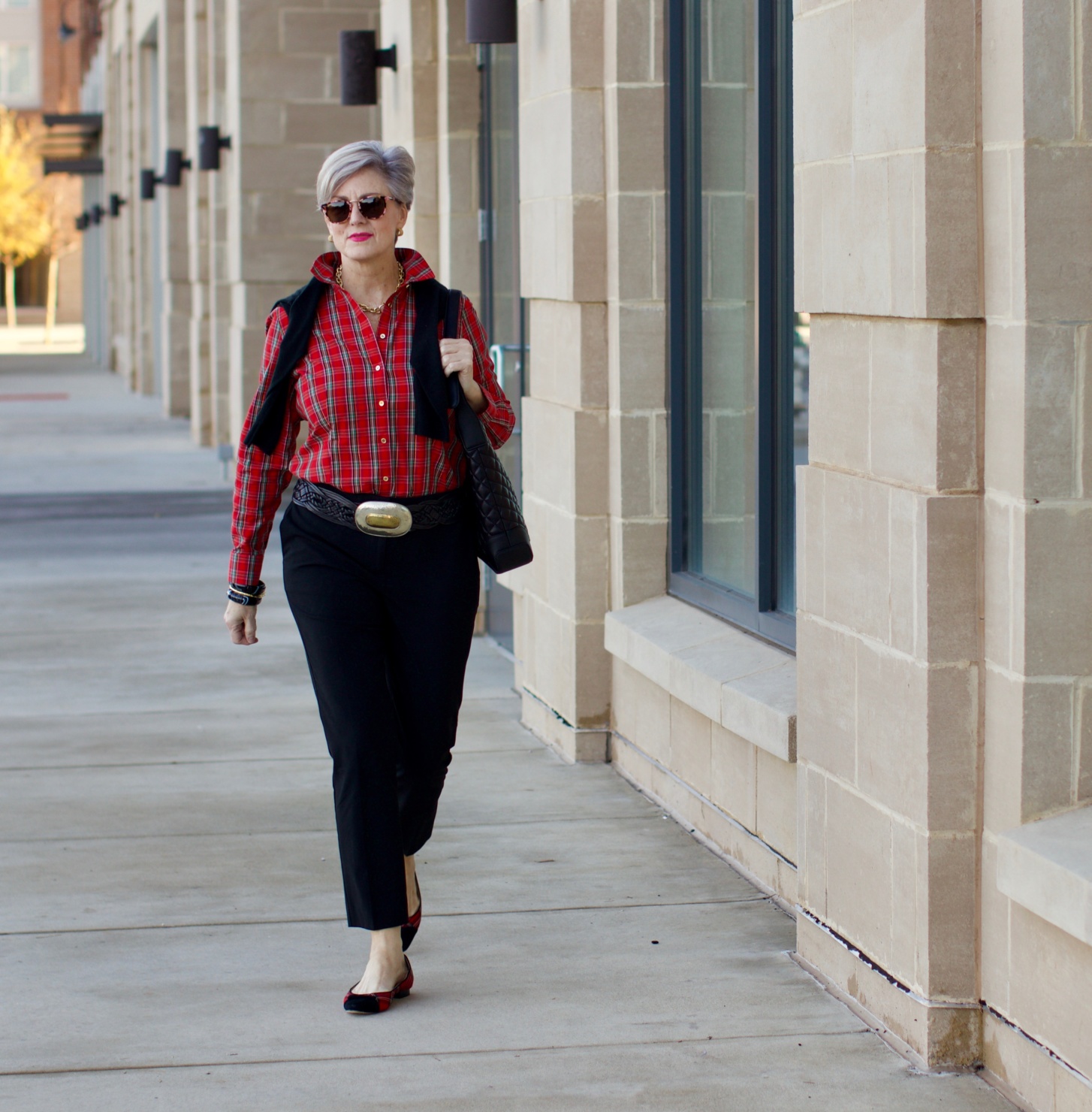 beth from Style at a Certain Age wears a tartan plaid shirt, black cropped ankle pants, tartan plaid flats, military coat and quilted hobo handbag.