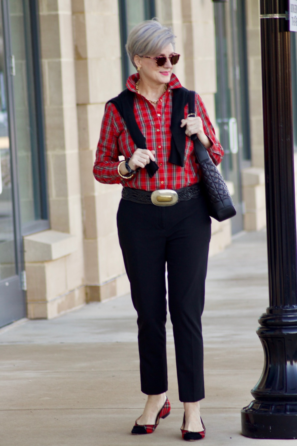 beth from Style at a Certain Age wears a tartan plaid shirt, black cropped ankle pants, tartan plaid flats, military coat and quilted hobo handbag.