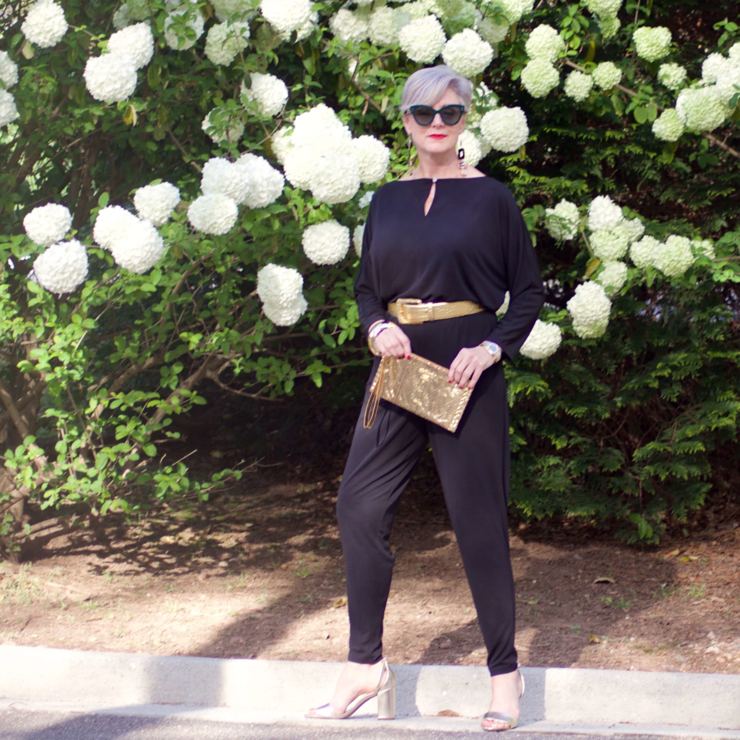 beth from Style at a Certain Age wears a black jumpsuit, gold sandals, gold belt, and gold clutch handbag