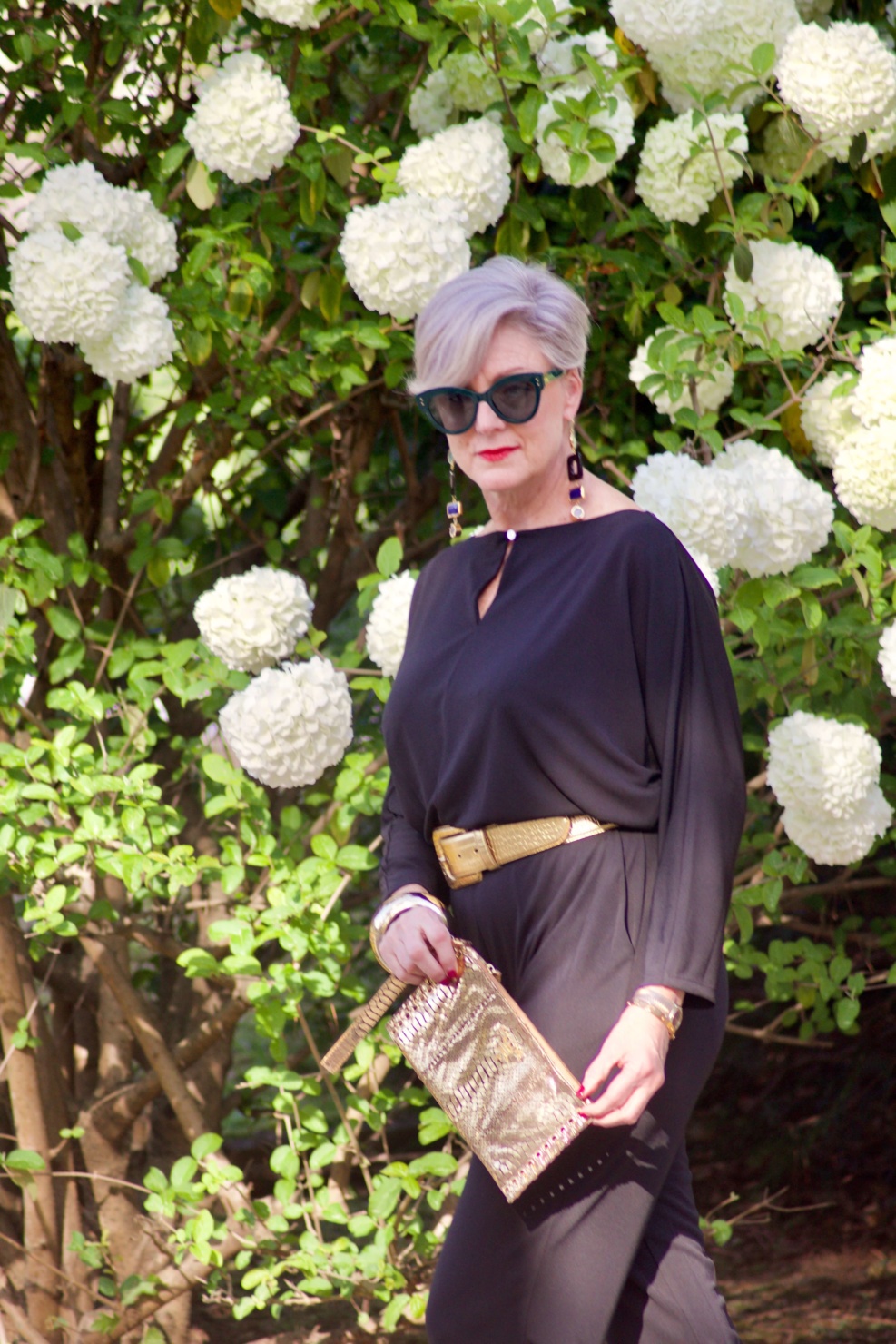 beth from Style at a Certain Age wears a black jumpsuit, gold sandals, gold belt, and gold clutch handbag