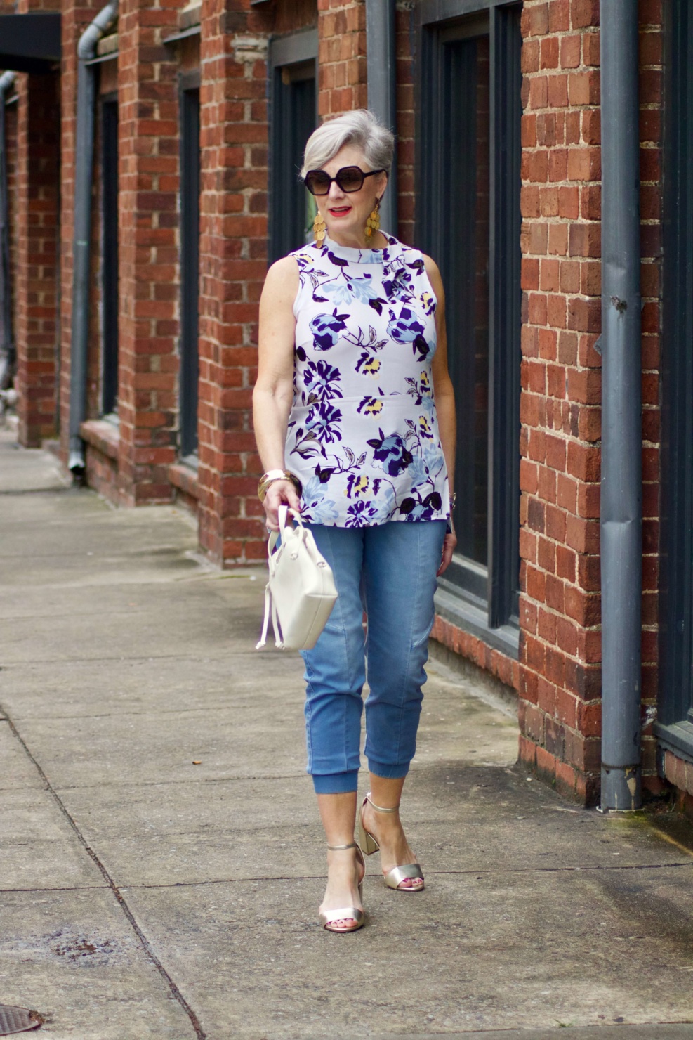 beth from Style at a Certain Age wears a floral top, denim joggers, gold metallic block sandals, and carries a drawstring crossbody handbag. Walmart we dress America